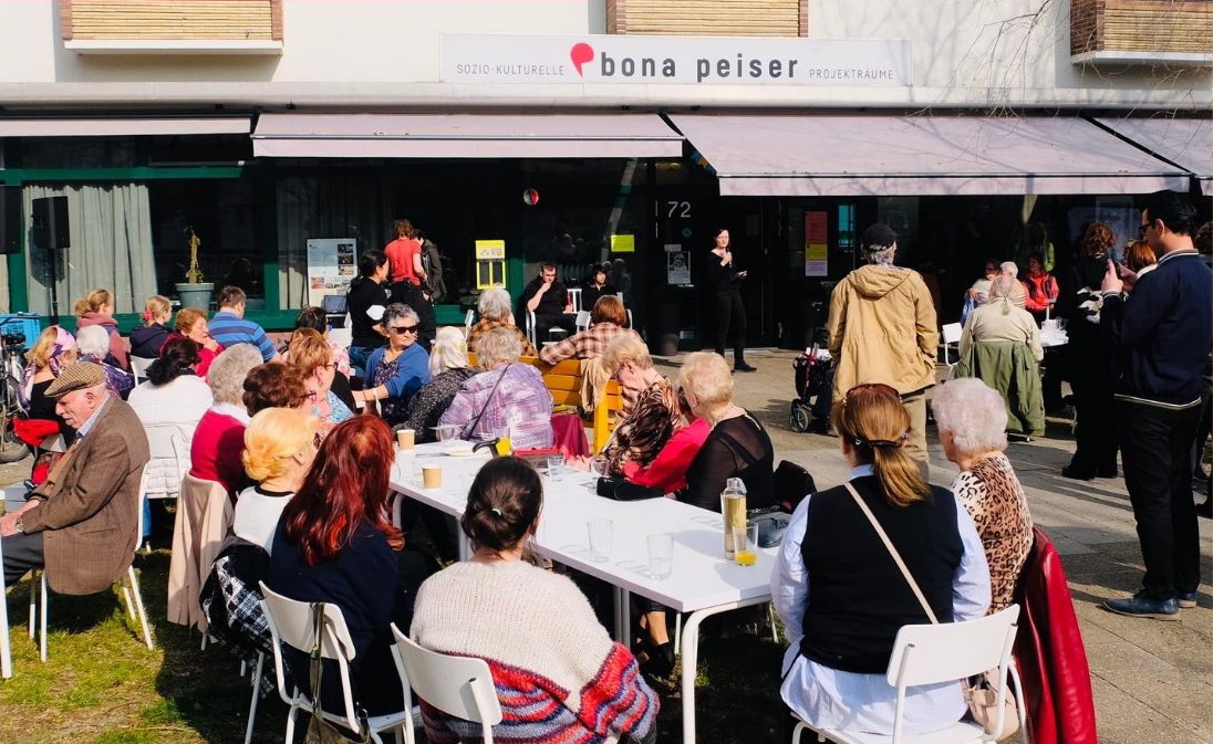 bunt gemischte Menschengruppe sitzt draußen an Tischen vor einem Haus mit Ladenlokal im Erdgeschoss