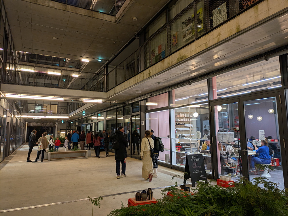 Betonterrasse bei Nacht, einige Menschen stehen in Gruppen beieinander, an der Seite ein hell erleuchtetes Atelier mit einigen Menschen darin