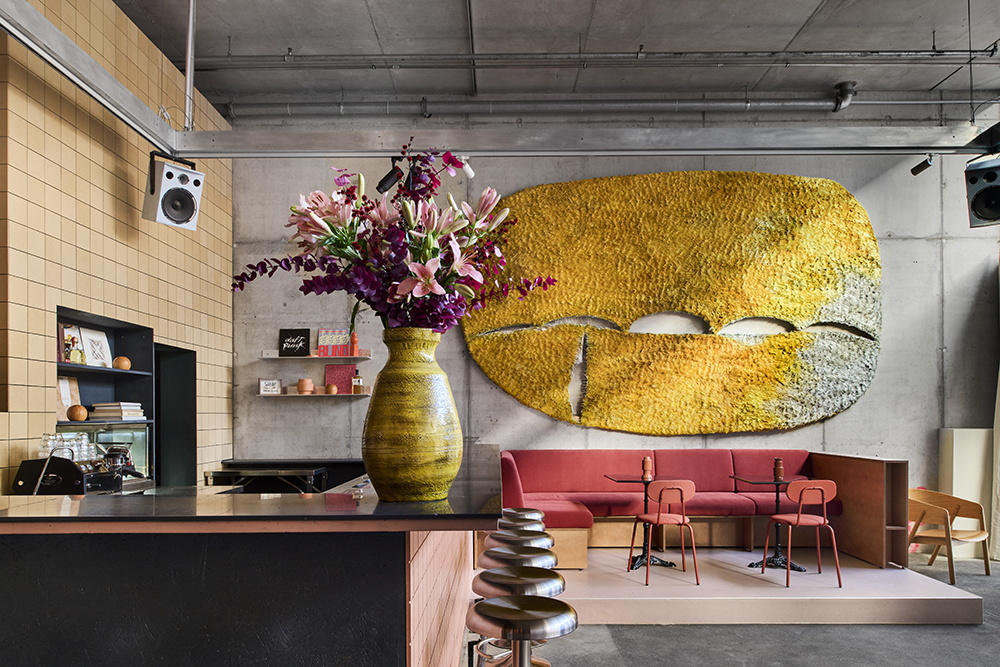 großer, heller Raum, auf der Ecke eines Tresens steht ein großer, üppiger Blumenstrauß in Rosatönen, im Hintergrund ein Koralle-farbenes Sofa vor einem großen gelben, teppich-artigen Kunstwerk