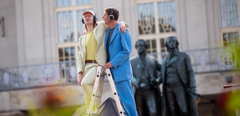 zwei junge Männer auf einer A Leiter vor dem Goethe und Schiller Denkmal am Theater Weimar, einer trägt einen gelben, der andere einen blauen Anzug, beide tragen große schwarze Kopfhörer, der Gelbe legt den Arm um den Blauen
