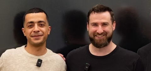 zwei junge Männer mit kurzen dunklen Haaren, der eine mit Vollbart, lächeln frontal in die Kamera
